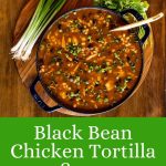 Vertical overhead photo of a pot of Mexican Black Bean Chicken Tortilla Soup on a wood platter surrounded by garnishes with a large word text title graphic at the bottom of the photo.