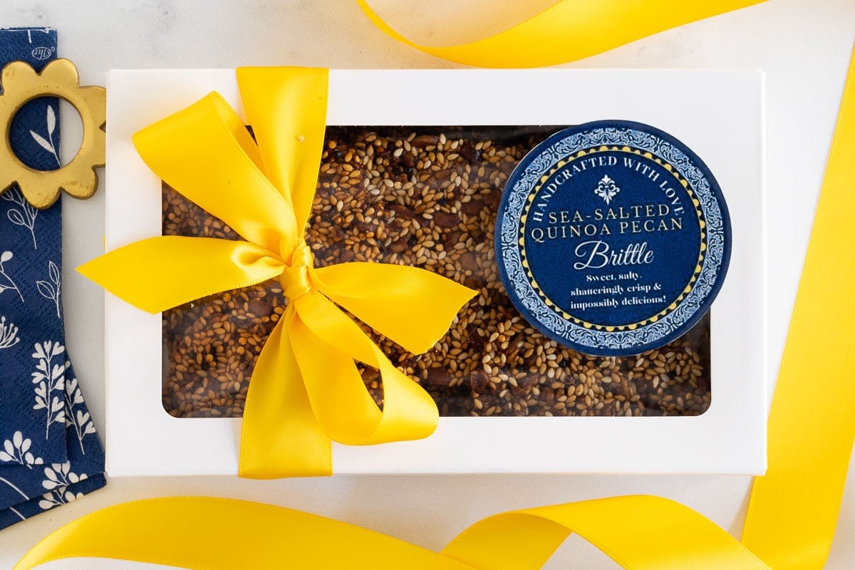 Horizontal overhead photo of a gift box filled with Sea-Salted Quinoa Pecan Brittle with a custom gift label and yellow ribbon.