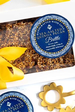 A photo of the Quinoa Pecan Brittle in a white box with a clear surface , decorated with a yellow ribbon bow and a blue and white colored round label.