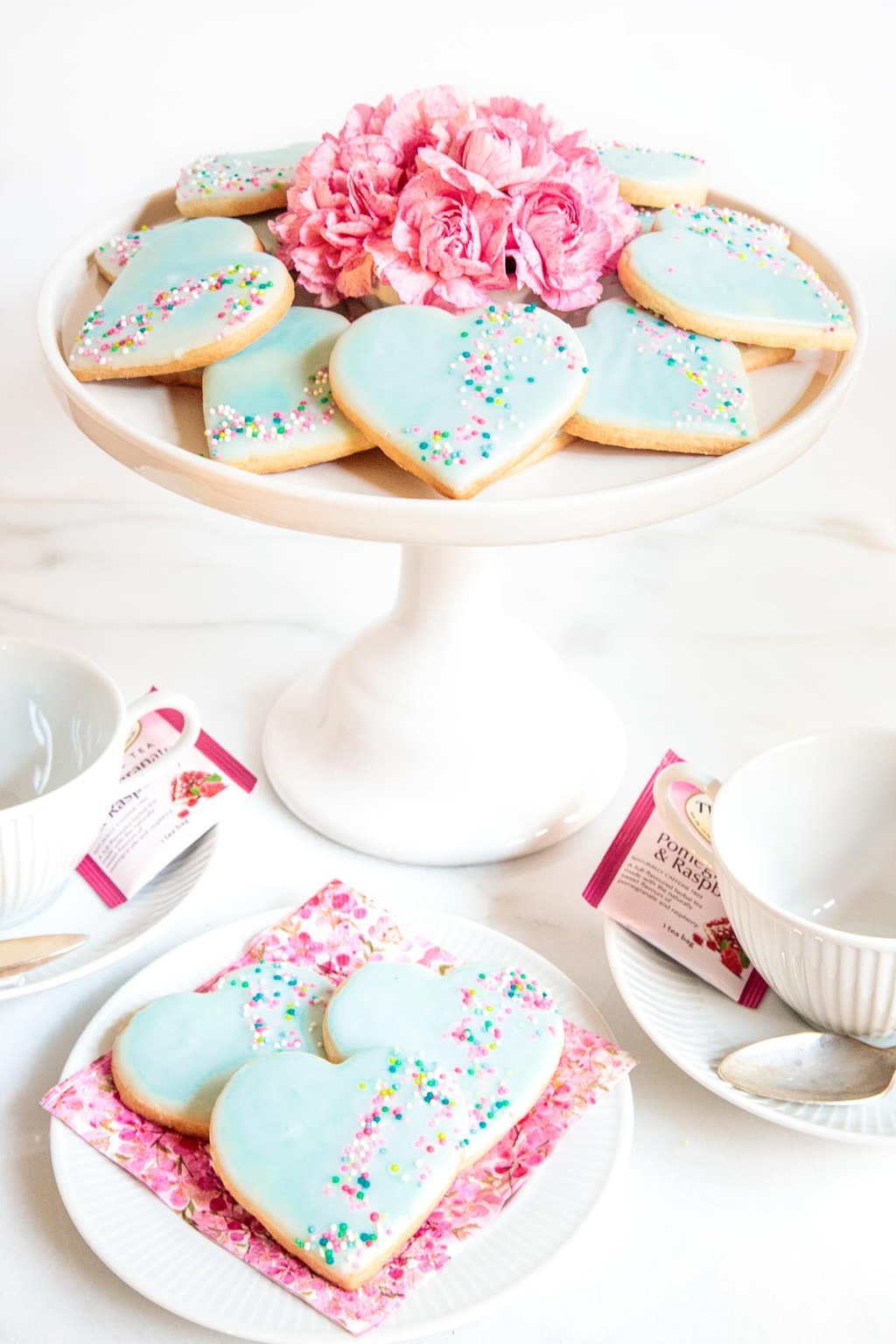 Vertical photo of a batch of Valentine Shortbread Cookies on a white pedestal serving plate decorated with flowers.