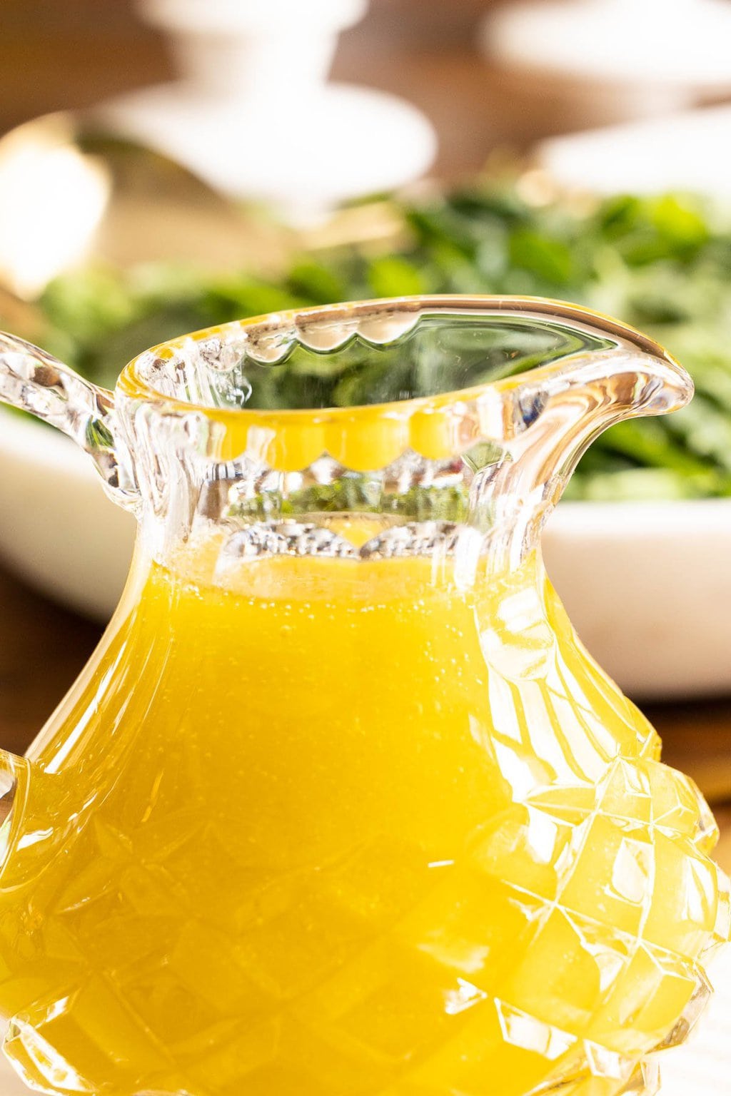 Vertical extreme closeup photo of a cut glass pitcher of Honey White Balsamic Dressing with a salad in the background.