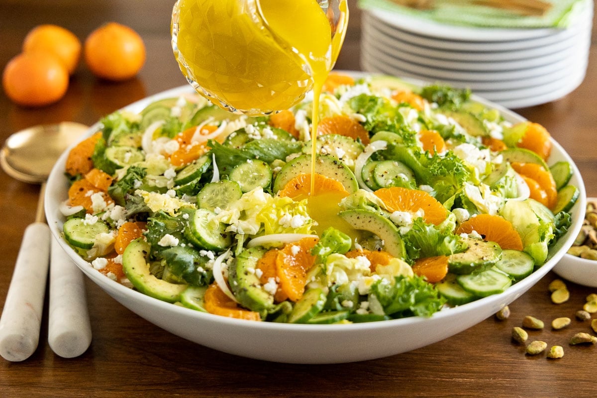 Horizontal photo of an Irish Flag Clementine Cucumber Salad in a large white serving bowl on a wood table with Honey White Balsamic Dressing being poured over the top.