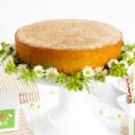 Horizontal photo of a Ridiculously Easy Mexican Churro Cake on a white pedestal cake plate surrounded with flowers.
