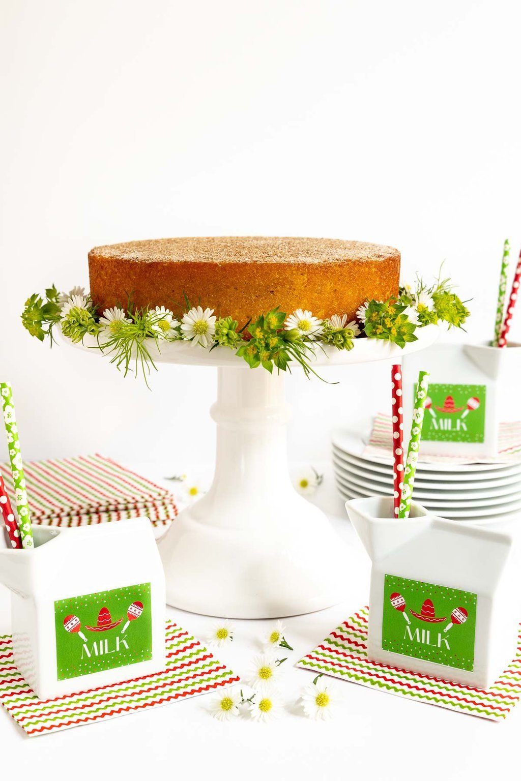Vertical photo of a Ridiculously Easy Mexican Churro Cake on a white pedestal cake plate decorated with flowers and surrounded by cartons of milk with straws.