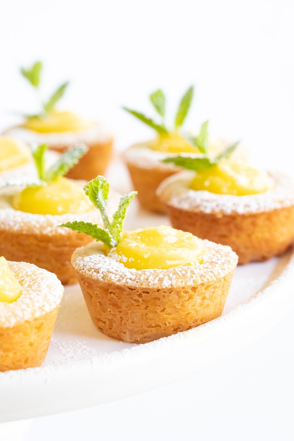 Vertical closeup photo of a batch of Lemon Curd Shortbread Tarts decorated with powdered sugar and a garnish of fresh mint leaves.
