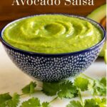 Vertical closeup photo of a bowl of Fresh Tomatillo Avocado Salsa surrounded by cilantro leaves and an avocado. A word title graphic is at the top of the photo.