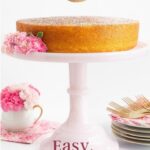 Vertical closeup photo of a person sifting powdered sugar on top of a French Butter Cake on a pale pink pedestal cake stand decorated with spring flowers.