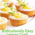 Vertical closeup photo of a batch of Ridiculously Easy Lemon Curd Tarts on a white pedestal platter sprinkled with powdered sugar and garnished with a sprig of fresh mint.