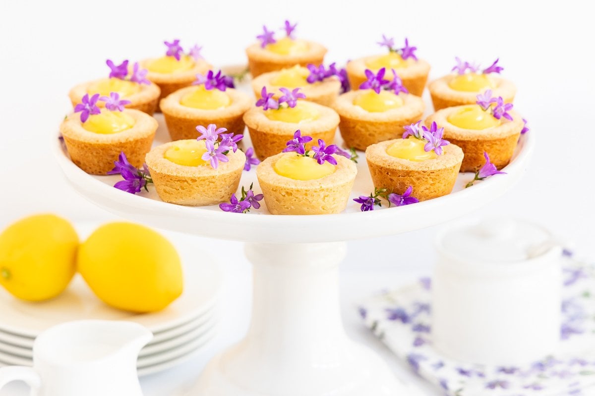Horizontal photo of a batch of Lemon Curd Shortbread Tarts garnished with edible flowers on a white pedestal serving plate.