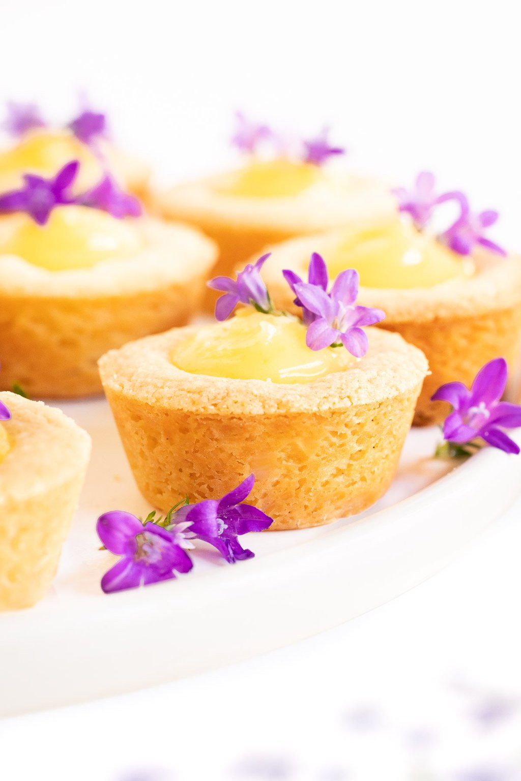 Vertical extreme closeup photo of a batch of Ridiculously Easy Lemon Curd Shortbread Tarts on a white pedestal serving stand garnished with purple flowers.