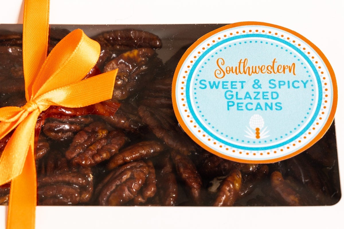Horizontal extreme closeup photo of a gift box of Southwest Sweet and Spicy Glazed Pecans decorated with a gold bow and custom gifting labels.