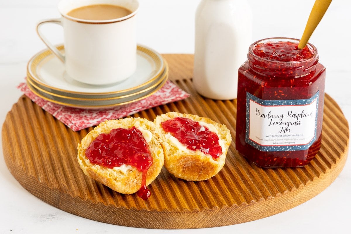 Horizontal photo of a jar of Strawberry Raspberry Lemongrass Jam with the jam spread on homemade rolls in the foreground.