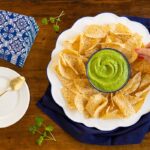 Horizontal overhead photo of a dish of Fresh Tomatillo Avocado Salsa surrounded by tortilla chips. A person is dipping a chip into the salsa.