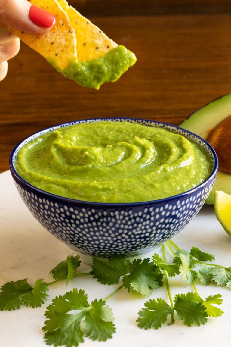 Vertical picture of Tomatillo Avocado Salsa in a blue and white bowl with a chip being dipped in the salsa.