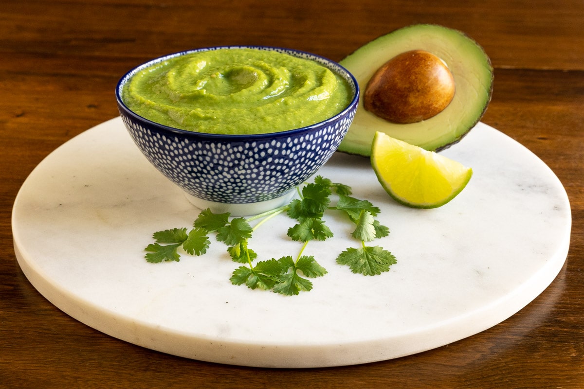 Horizontal photo of a dish of Fresh Tomatillo Avocado Salsa surrounded by an avocado, cilantro and a lime wedge on a round marble surface.