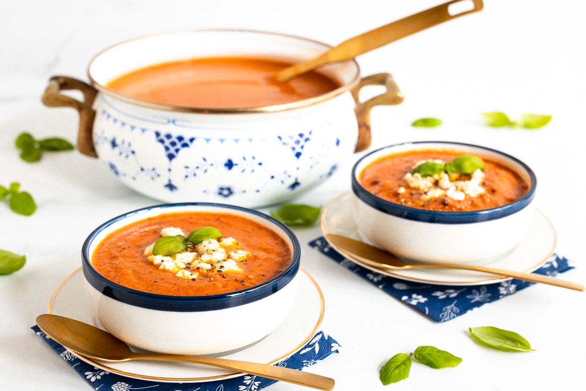 Horizontal photo of two serving bowls and a pot of White Bean Tomato Basil Soup surrounded by fresh basil leaves.