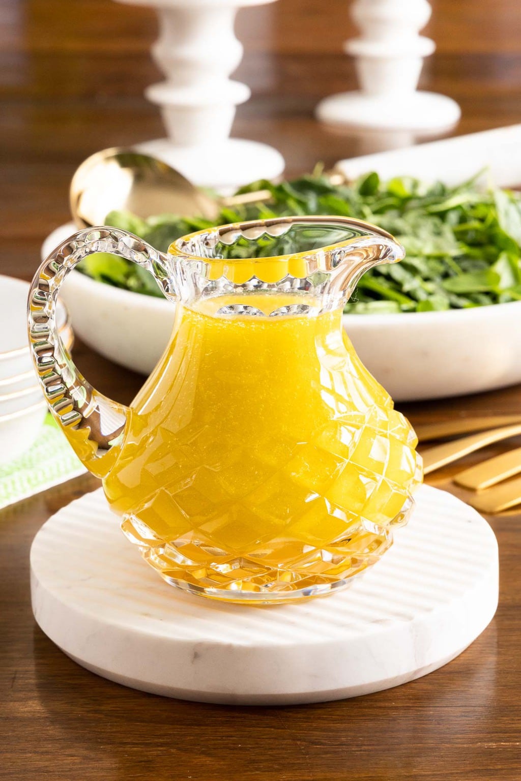 Vertical closeup photo of a cut glass pitcher filled with Honey White Balsamic Dressing on a round marble surface with a spinach salad in the background.