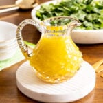 Horizontal photo of a cut glass pitcher filled with Honey White Balsamic Dressing on a wood table.