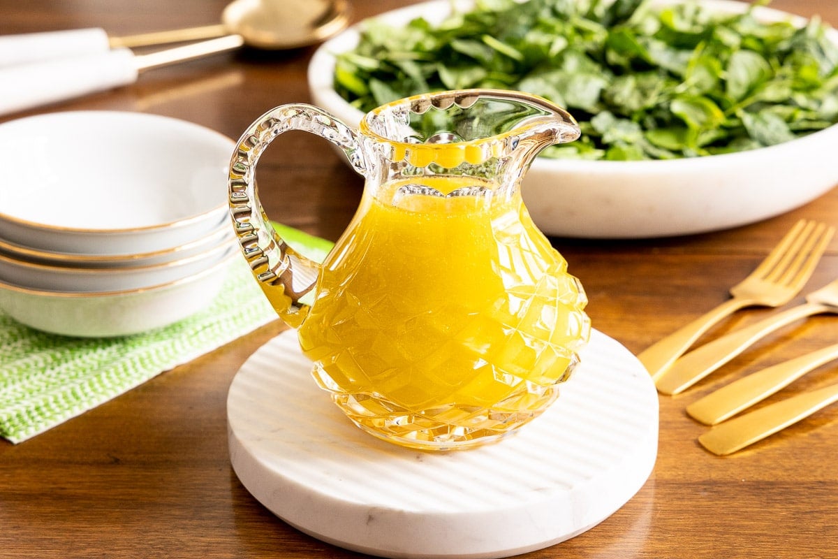 Horizontal photo of a cut glass pitcher of Honey White Balsamic Dressing on a small round marble surface with a spinach salad in the background.