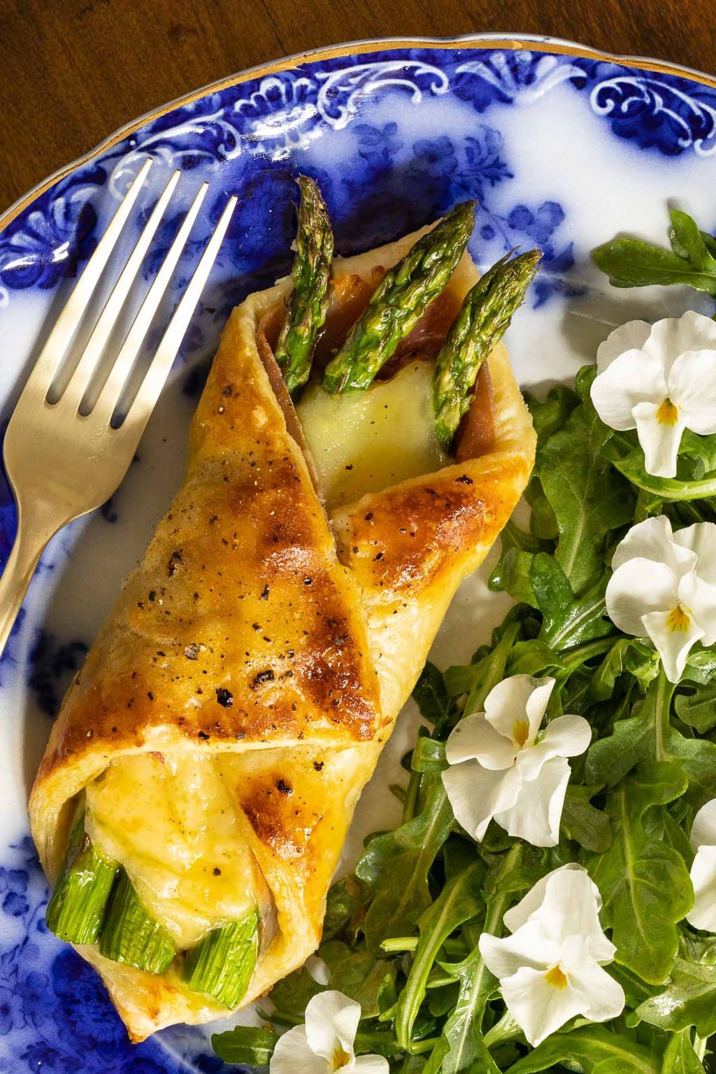 Vertical extreme closeup photo of a Honey-Glazed Asparagus Prosciutto Puff Pastry Wrap with an arugula side salad decorated with edible pansy flowers.
