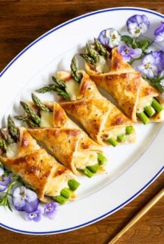 Horizontal overhead photo of a white serving plate of Honey-Glazed Asparagus Prosciutto Puff Pastry Wraps decorated with edible pansies.