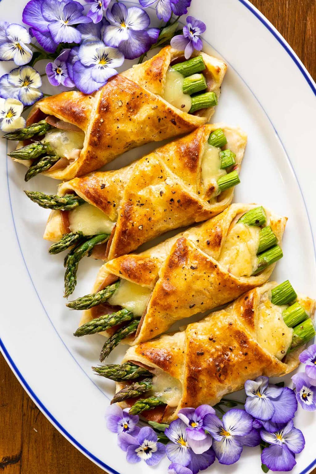 Vertical closeup overhead photo of an oval serving plate with Honey-Glazed Asparagus Prosciutto Puff Pastry Wraps garnished with edible pansy flowers.