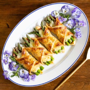 Horizontal overhead photo of a white serving plate of Honey-Glazed Asparagus Prosciutto Puff Pastry Wraps decorated with edible pansies.