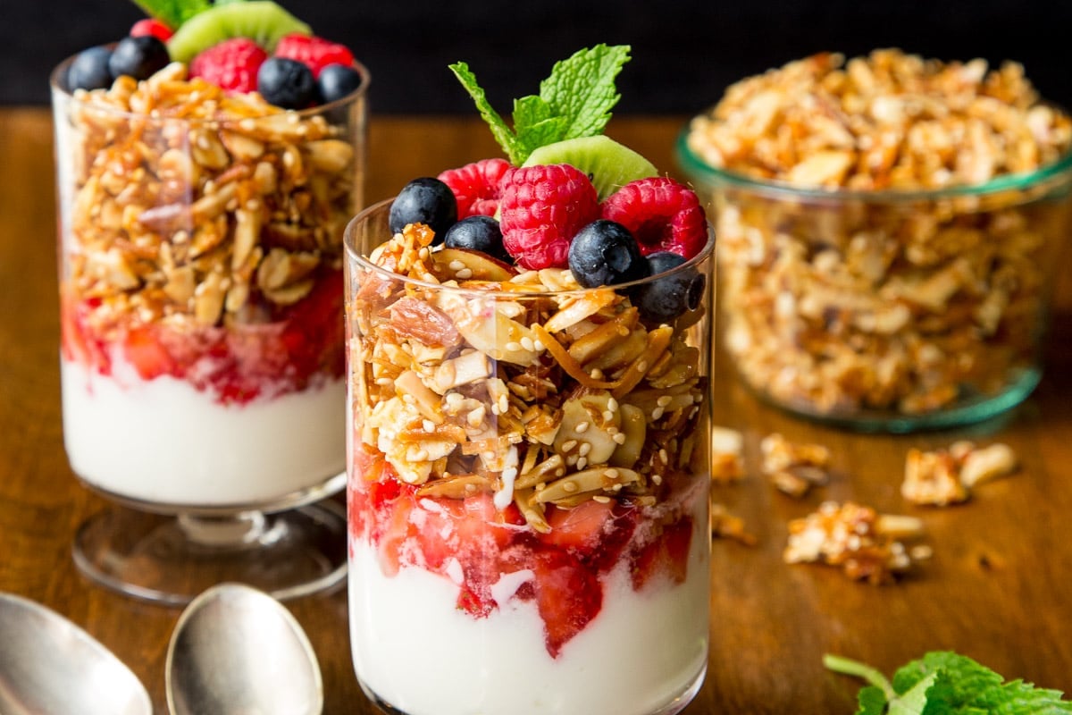 Horizontal closeup photo of several Double Almond Coconut Granola parfaits garnished with fresh fruit and mint leaves.