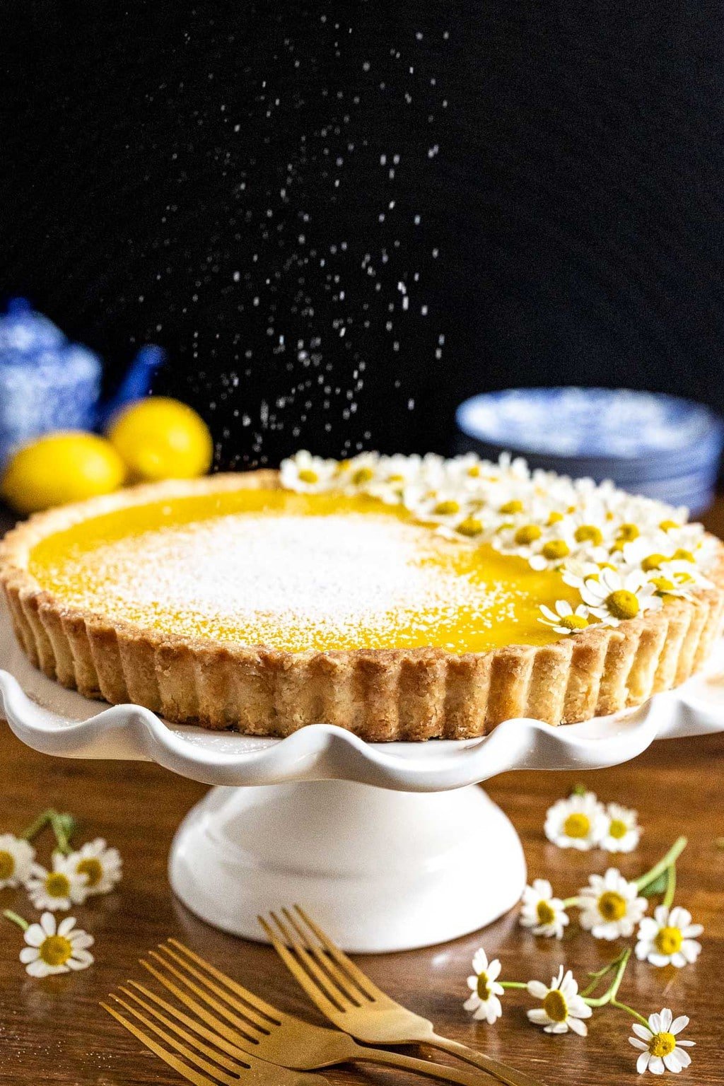 Vertical photo of powdered sugar floating down on a French Lemon Tart on a white pedestal serving plate surrounded and decorated with camomile flowers.