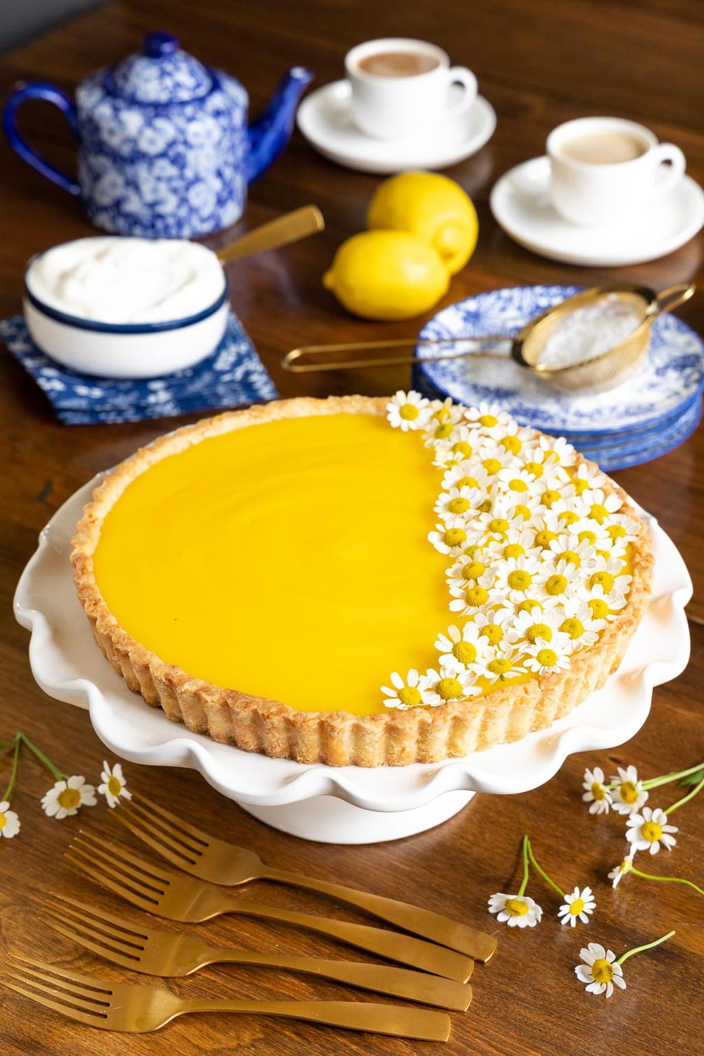 Vertical photo of French Lemon Tart decorated with camomile flowers on a white pedestal cake stand with cups of coffee in the background.