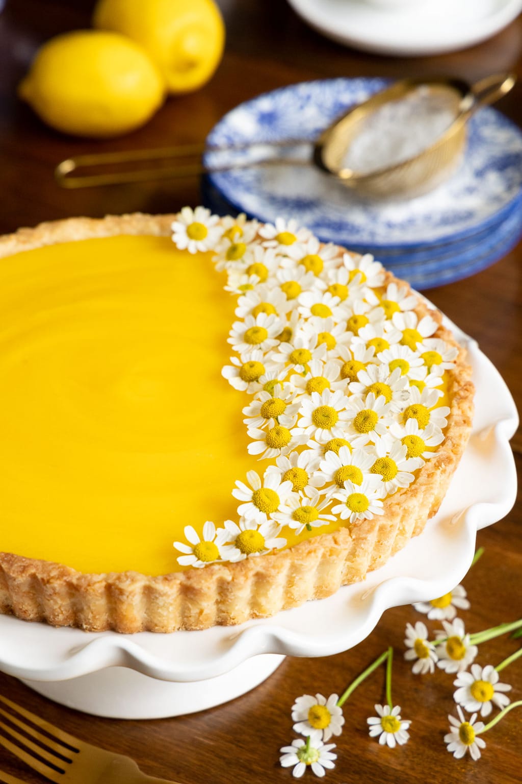 Vertical closeup photo of a French Lemon Tart on a white pedestal serving plate and decorated with edible chamomile flowers.