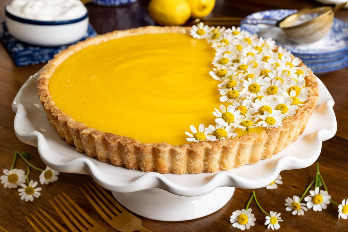 Horizontal closeup photo of a French Lemon Tart on a white pedestal serving plate and decorated with edible chamomile flowers.