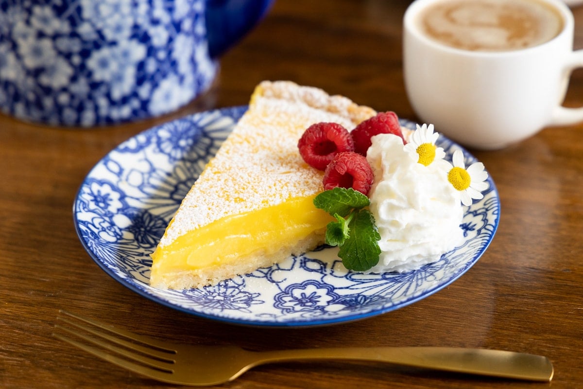 Horizontal photo of a slice of a French Lemon Tart garnished with fresh raspberries, mint leaves, a dollop of whipped cream and camomile flowers.