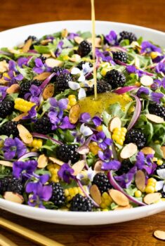 Horizontal closeup photo of a Fresh Corn Blackberry Arugula Salad decorated with flowers on a wood table.