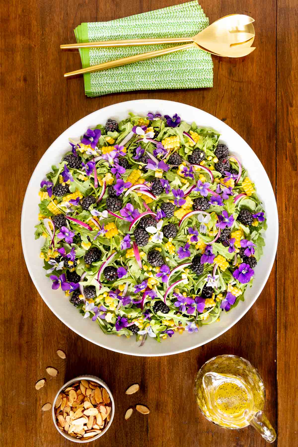 Vertical overhead photo of a Blackberry Arugula Salad in a white serving bowl and garnished with sliced almonds and purple edible wild flowers.