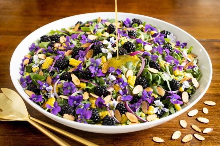 Horizontal closeup photo of a Fresh Corn Blackberry Arugula Salad decorated with flowers on a wood table.