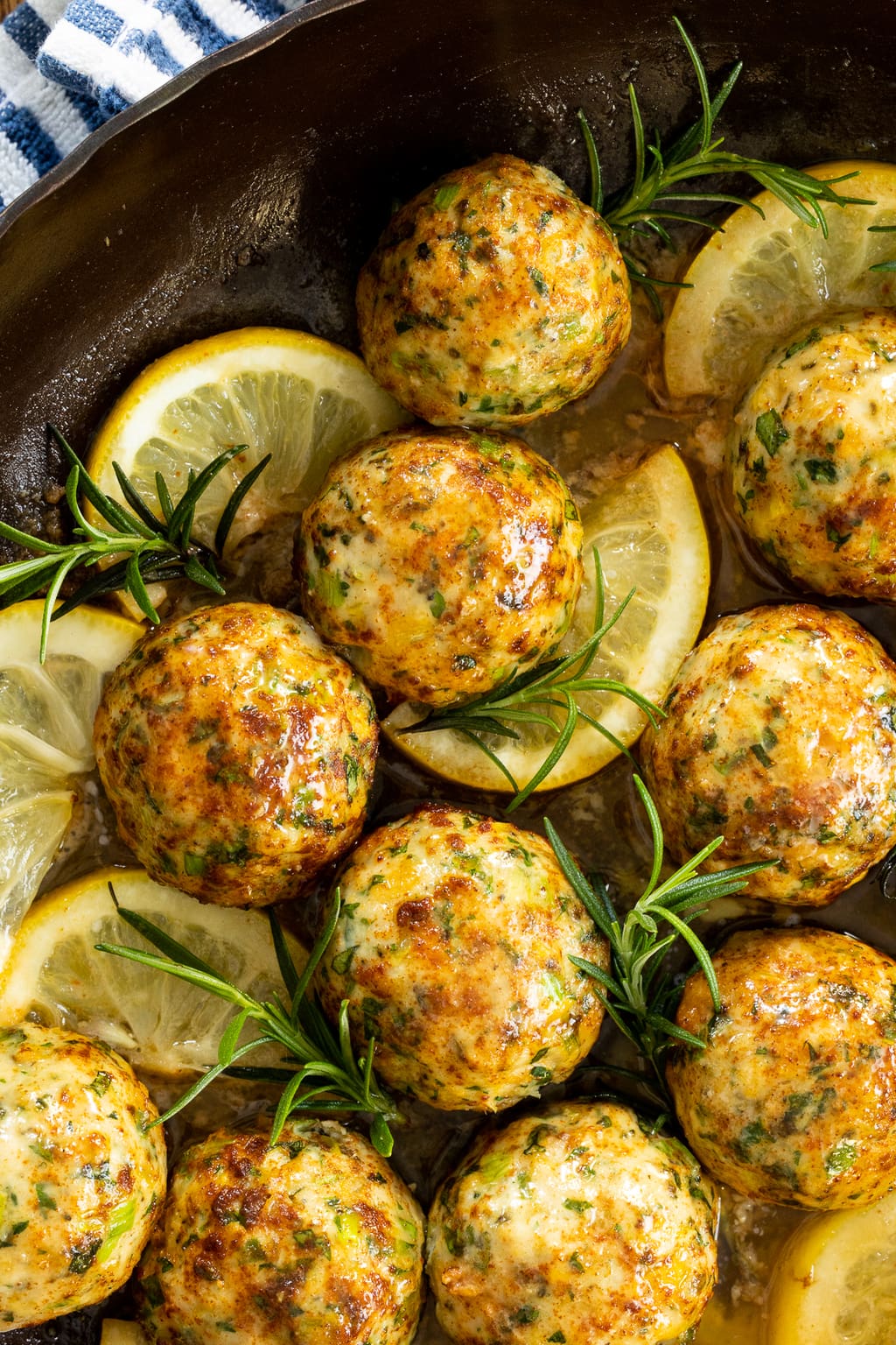 Extreme closeup photo of a batch of Fresh Herb Chicken Meatballs garnished with sprigs of fresh rosemary leaves and lemon wedges.