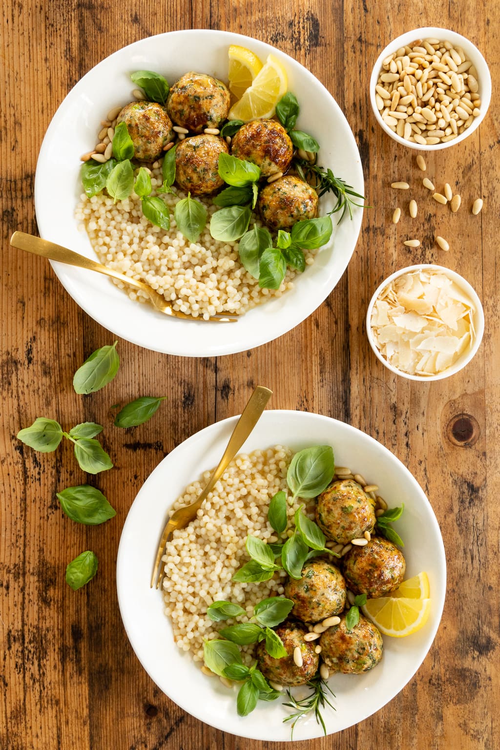 Vertical overhead photo of two white serving bowls filled with Fresh Herb Chicken Meatballs, couscous and fresh basil leaves with mini bowls of shaved Parmesan cheese and pine nuts nearby.
