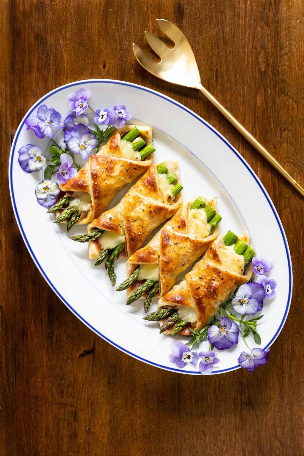 Vertical overhead photo of an oval serving plate with Honey-Glazed Asparagus Prosciutto Puff Pastry Wraps decorated with edible pansy flowers on a wood table.