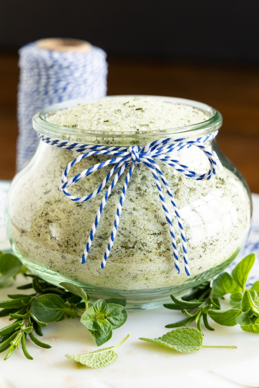 Vertical closeup photo of a glass Weck jar filled with Tuscan Herbed Sea Salt (Sale alle Erbe) and surrounded by fresh herbs.