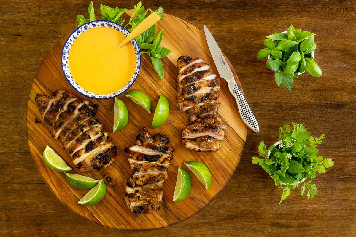 Overhead horizontal photo of Grilled Chicken Thighs with Coconut Chili Lime Sauce on a round wood platter garnished with fresh herbs and lime wedges.