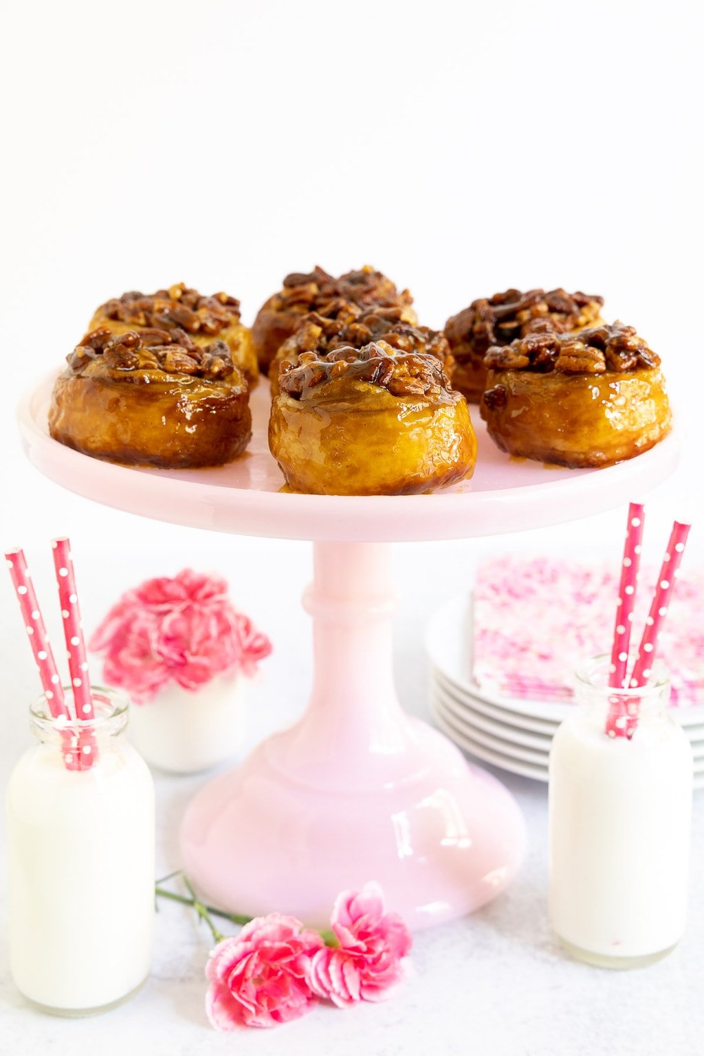 Vertical photo of a batch of Ridiculously Easy Sticky Pecan Rolls on a pale pink pedestal serving plate surrounded by jars of milk and pink straws.