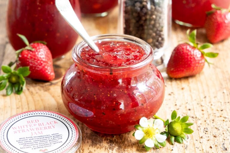 Jar of homemade strawberry balsamic black pepper jam with spoon, surrounded by fresh strawberries, black pepper grinder, and additional jars of jam on a wooden surface.