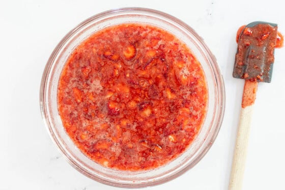 Overhead view of chopped strawberries mixed with sugar in a glass bowl, resting before making homemade strawberry freezer jam, with a spatula nearby.