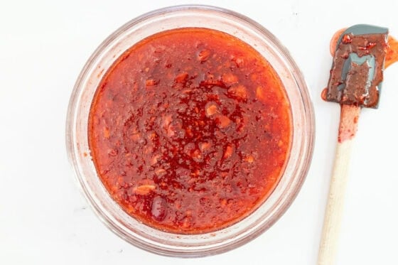 Overhead view of strawberry freezer jam mixture in a glass bowl after thickening, with a spatula beside the bowl.