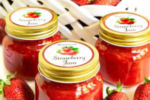 Three jars of homemade strawberry freezer jam with gold lids and strawberry labels, surrounded by fresh strawberries with a basket of berries in the background.