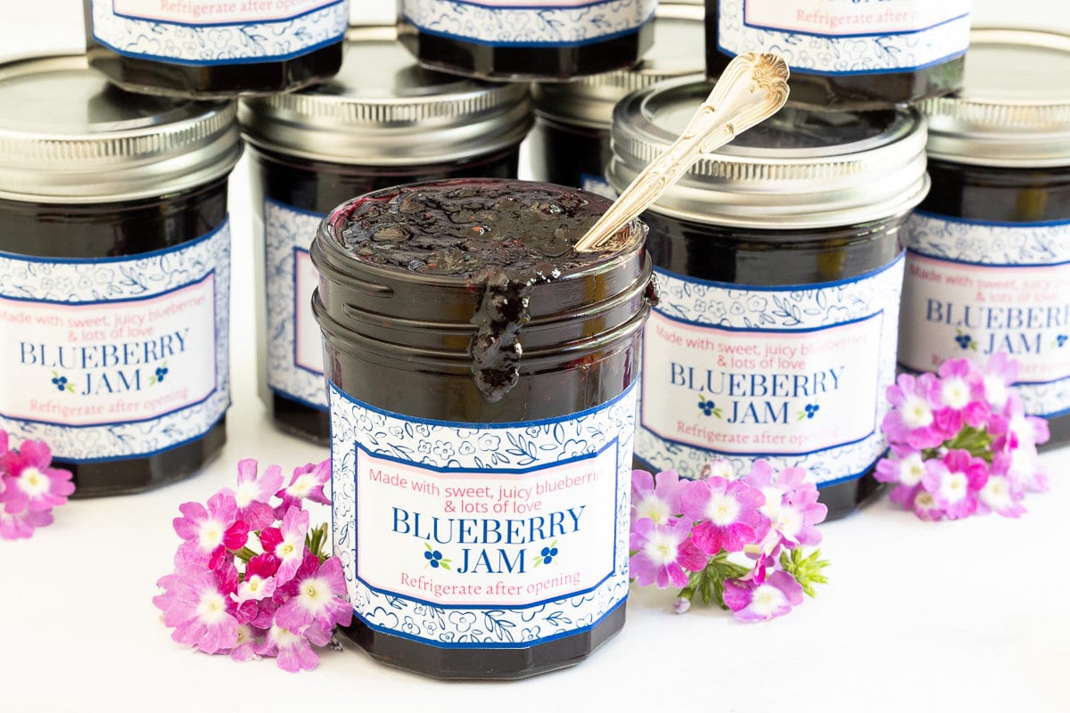 Horizontal closeup photo of jars of Easy 30-Minute Blueberry Jam with custom gift labels and surrounded by fresh flowers.