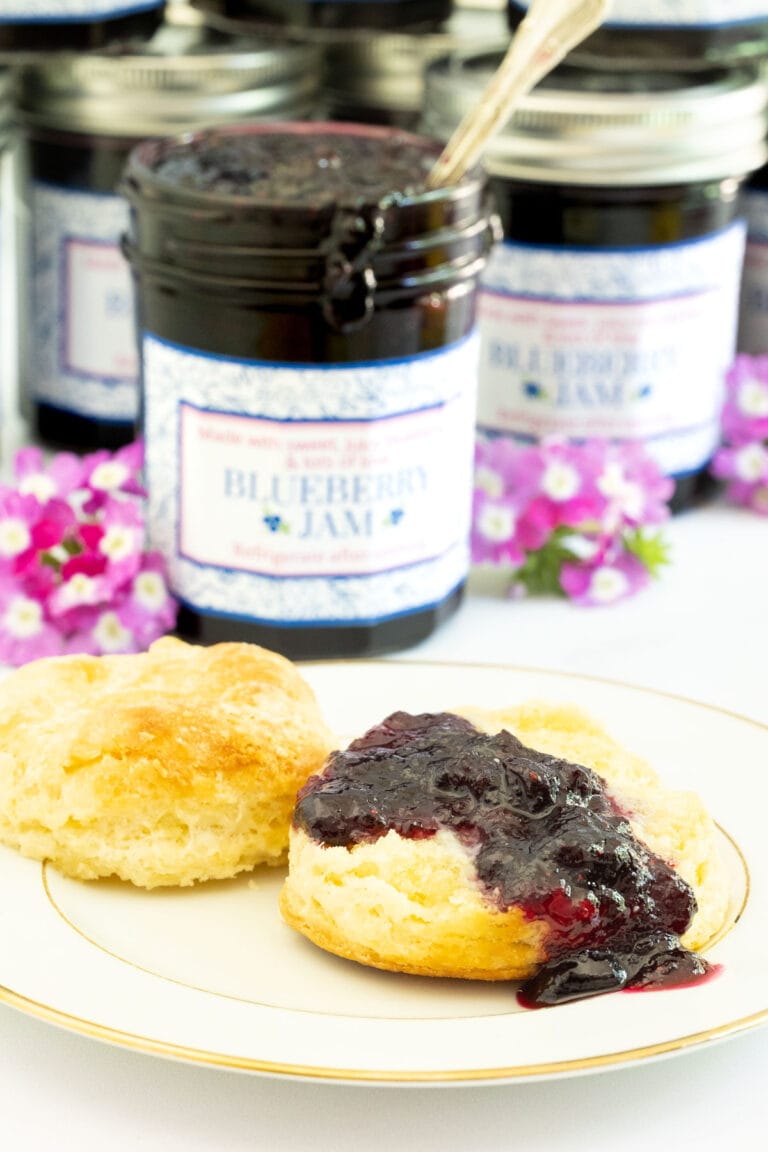 Vertical picture of Blueberry Jam in glass jars in the background and a scone with jam in the foreground