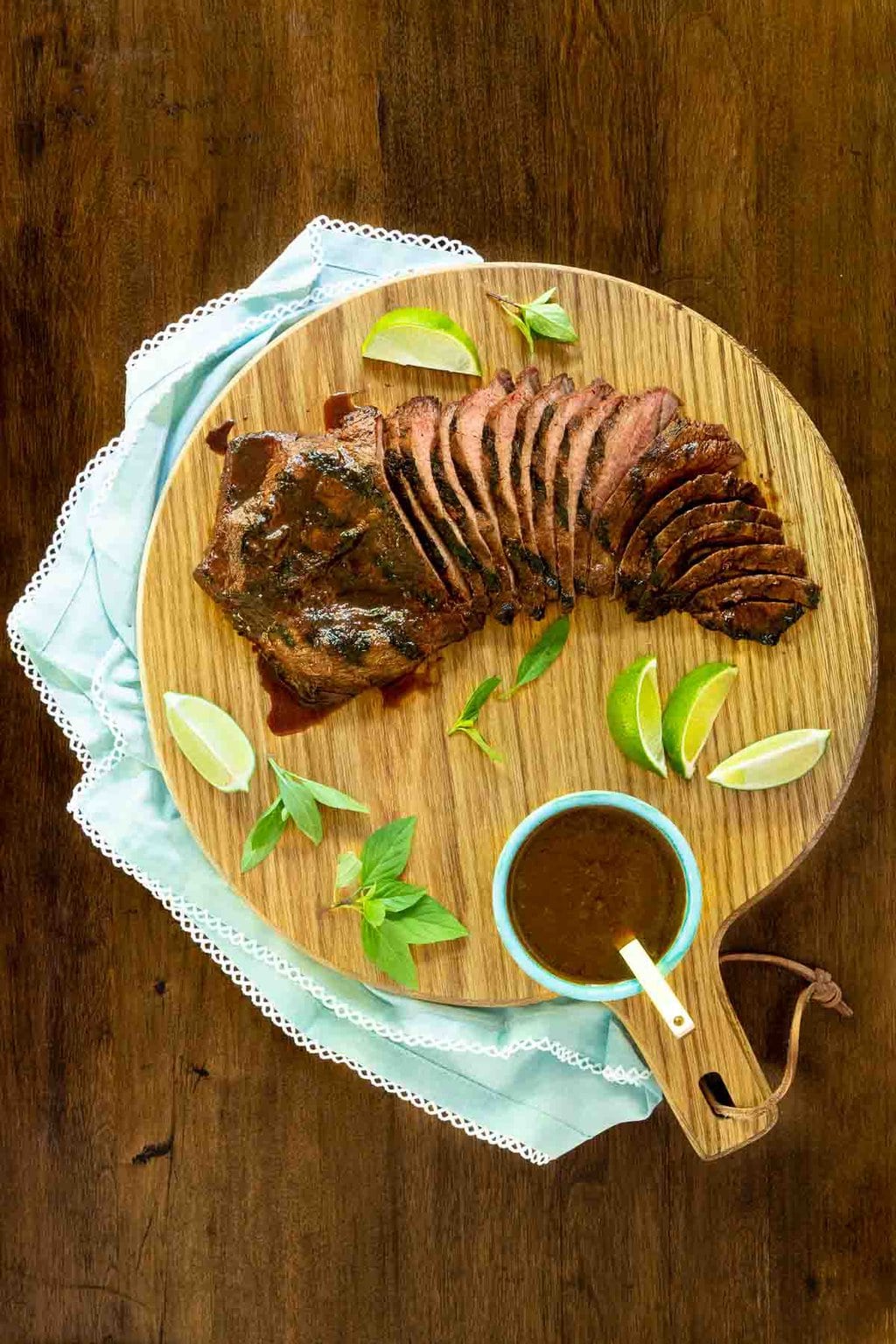 Vertical overhead photo of Steve's Grilled Asian Steak on a round wood cutting board with lime wedges, fresh herbs and a bowl of sauce.
