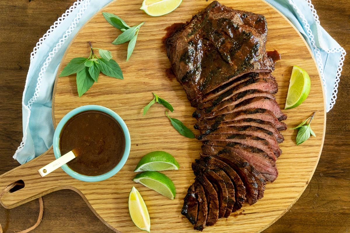 Horizontal overhead photo of Steve's Grilled Asian Steak on a wood cutting board surrounded by lime wedges and fresh herbs.